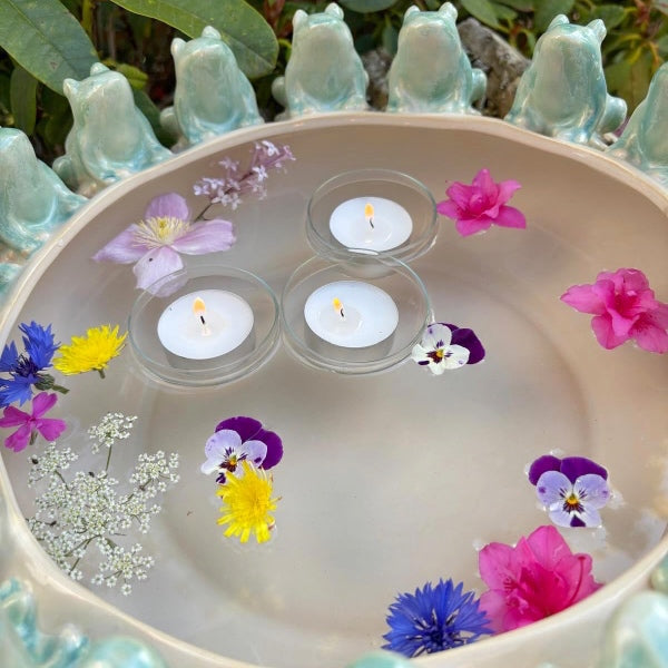 Stor keramisk skål med frømotiv fyldt med vand, flydende blomster og et tændt flydelys - dekorativ opstilling på bord.